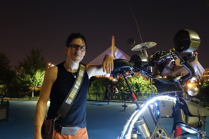 Man adorned with medals smiling in while leaning against a tallbike with the Boston skyline in the background