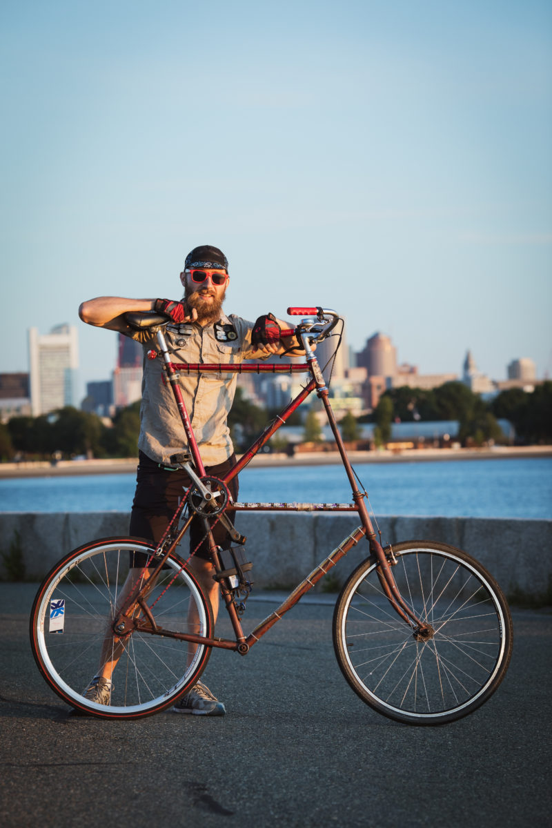 A bearded gentleman in dayglow orange sunglasses and a cycling cap proudly stands behind his tallbike with the Boston Skyline in the background..