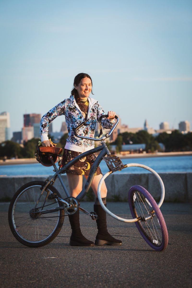 A proudly smiling your woman poses with her handbuilt chopper, with a fork that has outward curving tubes to form a a white circle, with the Boston Skyline in the background..