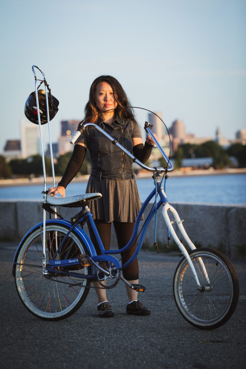 A young woman with a denim jacket, black leggings and arm warmers poses with her elegant home-made chopper, with the Boston Skyline in the background..